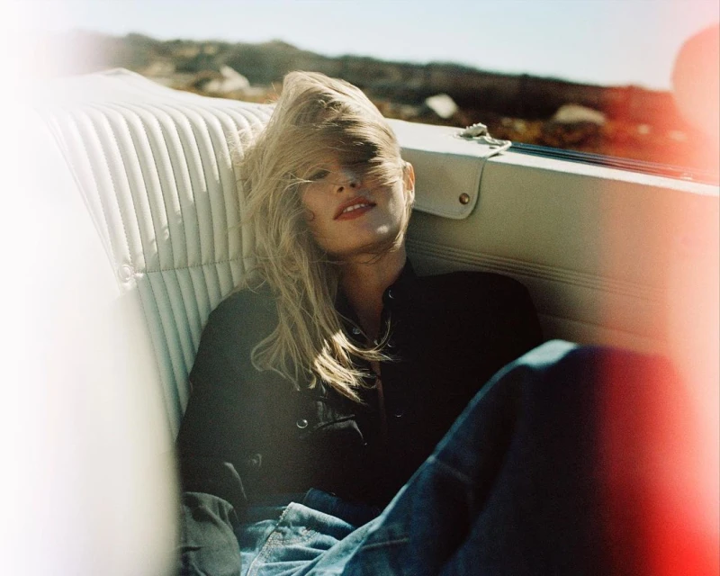 A woman with long blonde hair reclines in the white leather interior of a convertible car on a sunny day with a desert backdrop.