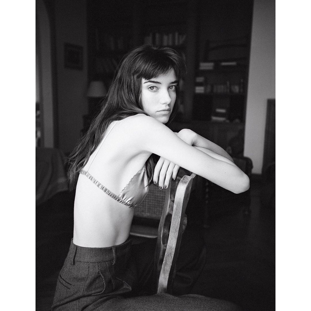 Monochrome portrait of a woman with dark hair sitting on a wooden chair in a room with a bookshelf in the background.