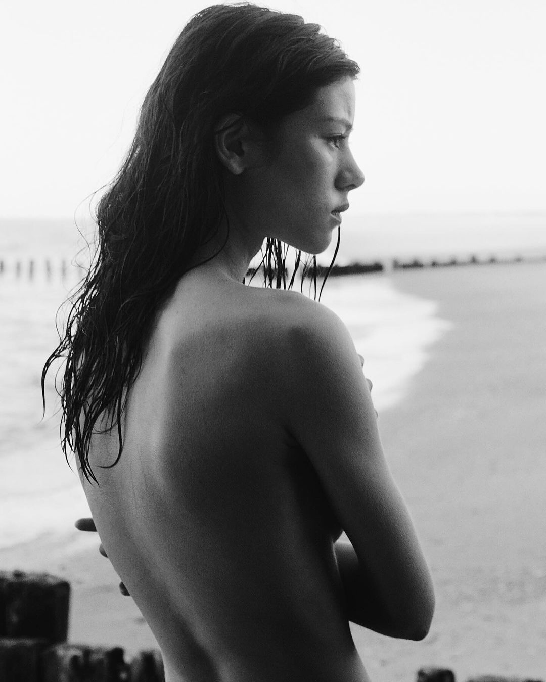 Black and white portrait of a young woman with wet hair on a beach with timber groynes, looking away with a contemplative expression.