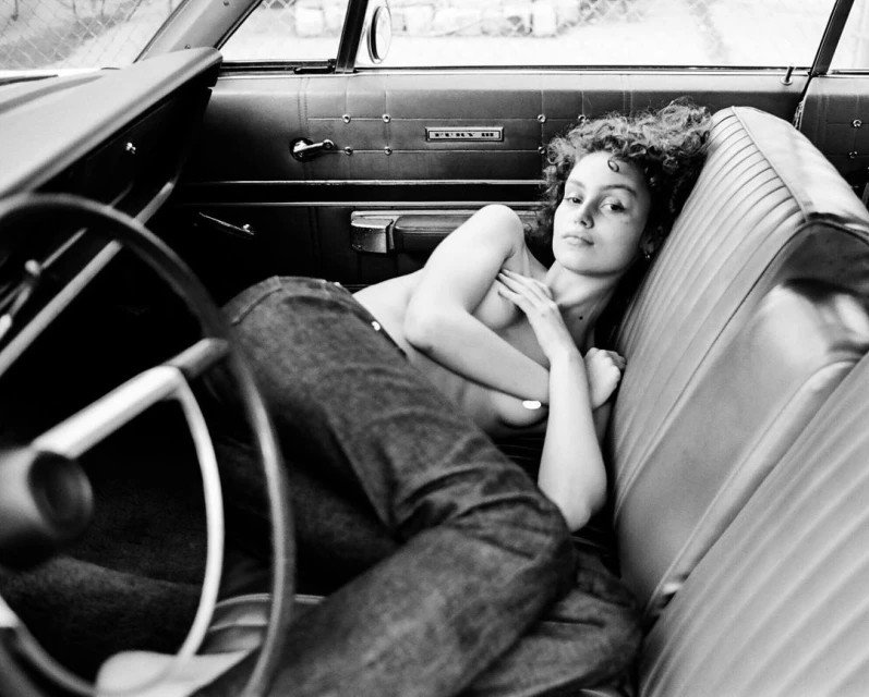 Monochrome portrait of a curly-haired woman reclining in the driver's seat of a vintage car, her chest partially covered by her arms.