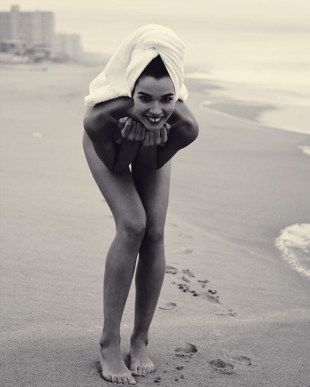 Monochrome portrait of smiling woman with white towel on head standing on sandy beach by ocean waves