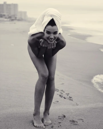 Monochrome portrait of smiling woman with white towel on head standing on sandy beach by ocean waves