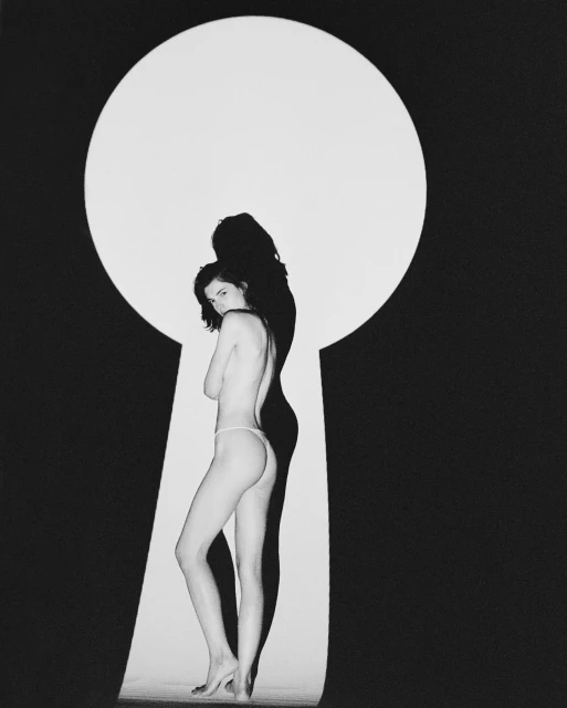 Adult female model stands in a photo studio in front of a keyhole-shaped light, posing in black and white with partial nudity.