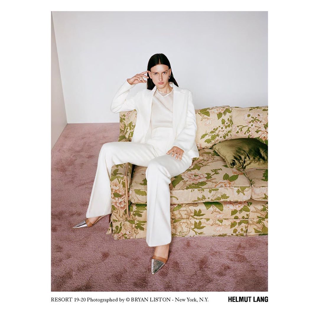 Elegant woman in a white suit poses on a floral couch in a studio with pink carpet, striking a sophisticated pose and fashionable look.