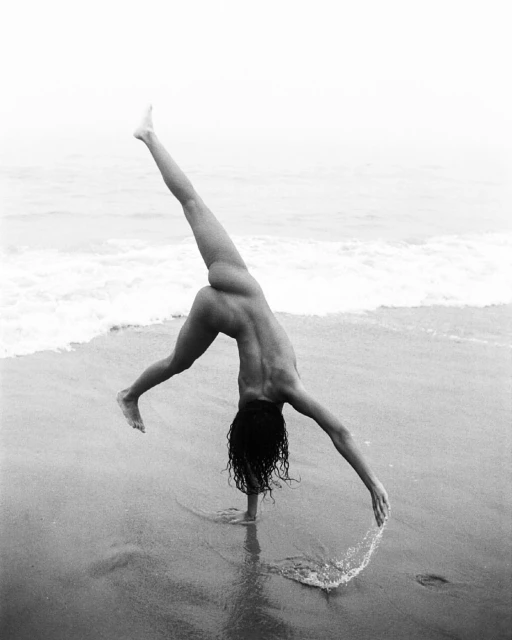 Nude adult female balances in cartwheel on wet sandy beach at ocean's edge; gray-scale, outdoor scene, overcast sky.