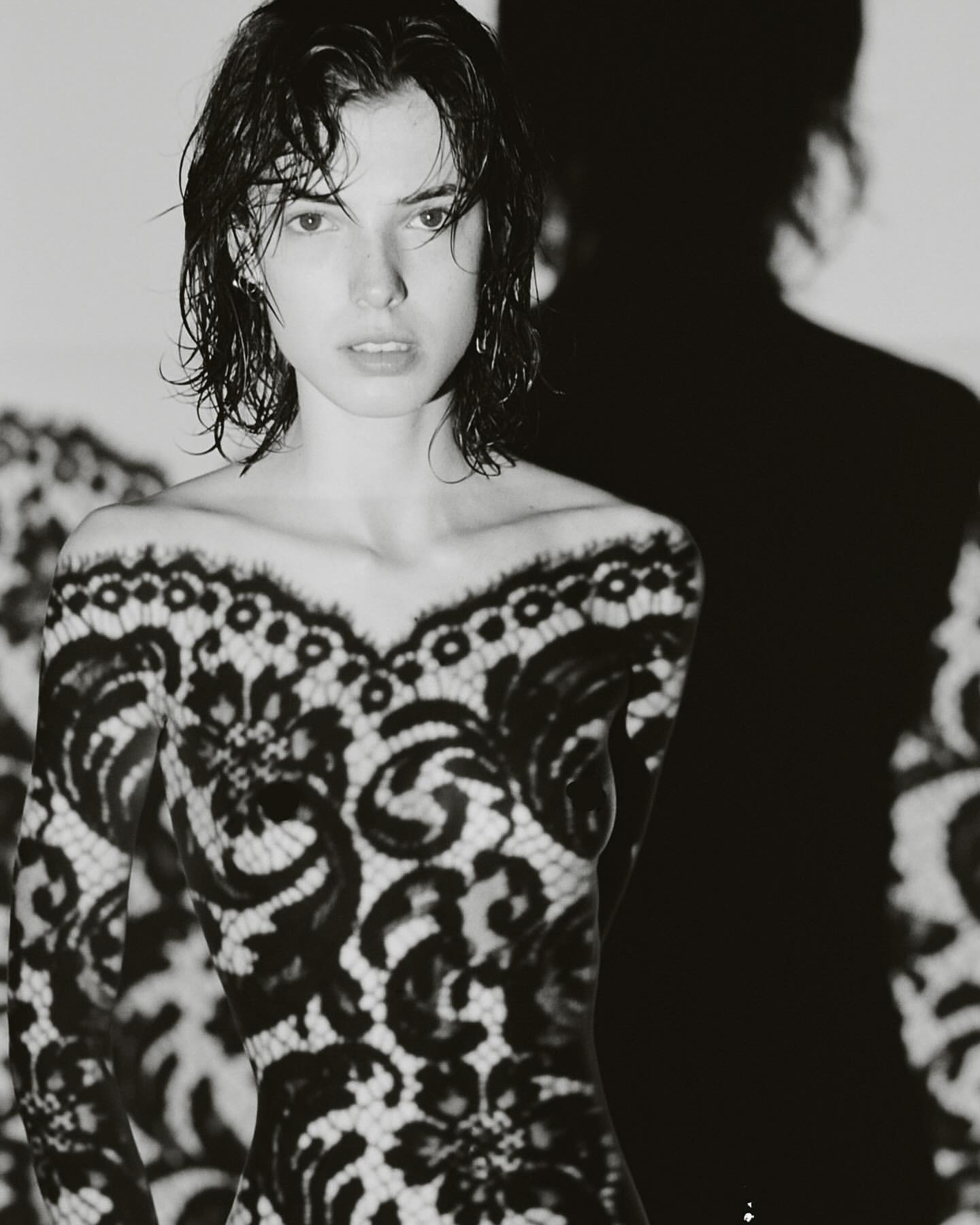 Black and white portrait of a woman with wet hair wearing a lace top against a white wall with a harsh shadow.