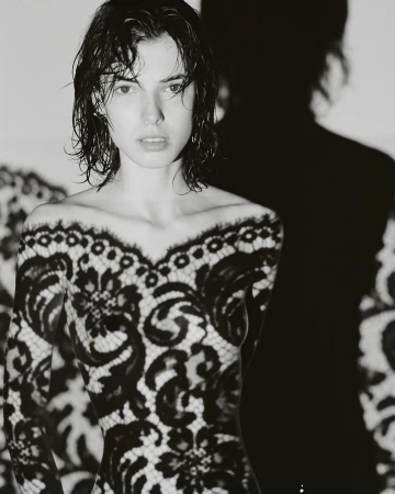 Black and white portrait of a woman with wet hair wearing a lace top against a white wall with a harsh shadow.