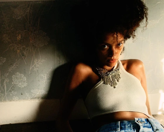 Woman in white halter top and rhinestone necklace leaning against a patterned wallpaper wall