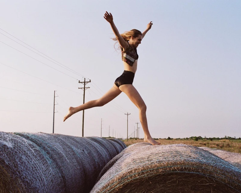 A woman in athletic wear leaps across a row of hay bales in a rural field under a clear sky, captured in a bright, natural light.