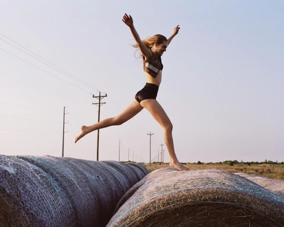 A woman in athletic wear leaps across a row of hay bales in a rural field under a clear sky, captured in a bright, natural light.