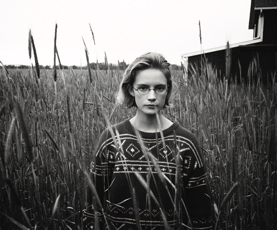 Black and white portrait of a young woman with glasses standing in a field of tall grass, wearing a patterned sweater with a house in the background.