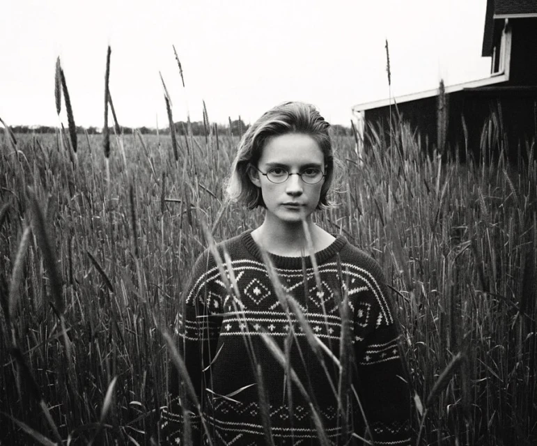 Black and white portrait of a young woman with glasses standing in a field of tall grass, wearing a patterned sweater with a house in the background.