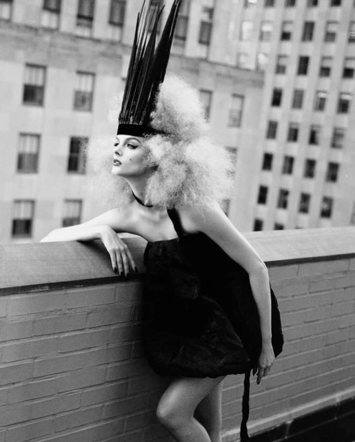 Black and white fashion portrait of a woman in a dark dress and elaborate feathered headdress, posing on a rooftop with city buildings behind.