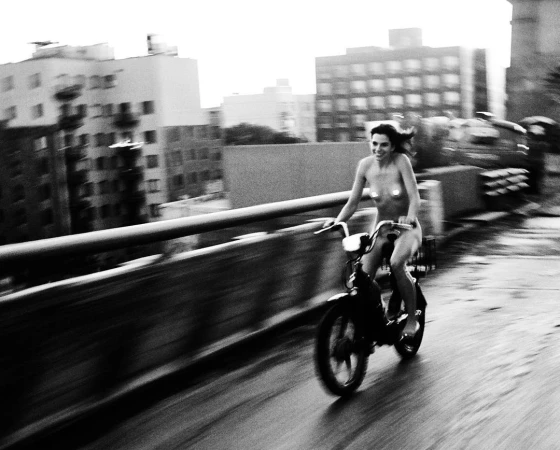 Monochrome action shot of a smiling woman riding a bicycle across a bridge with city buildings in the background.