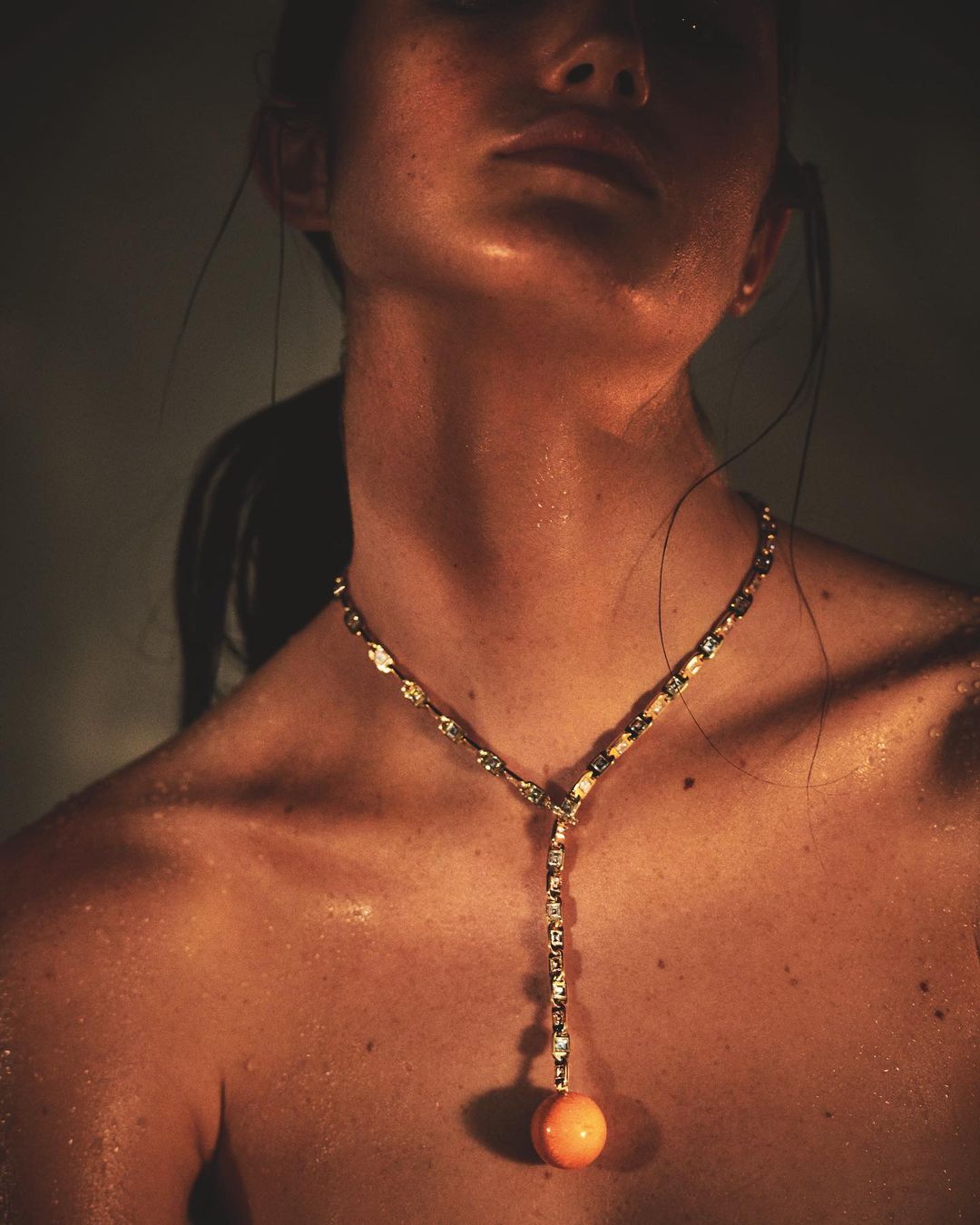 Close-up studio shot of an adult woman with sweaty skin, wearing a jeweled necklace with an orange spherical pendant.