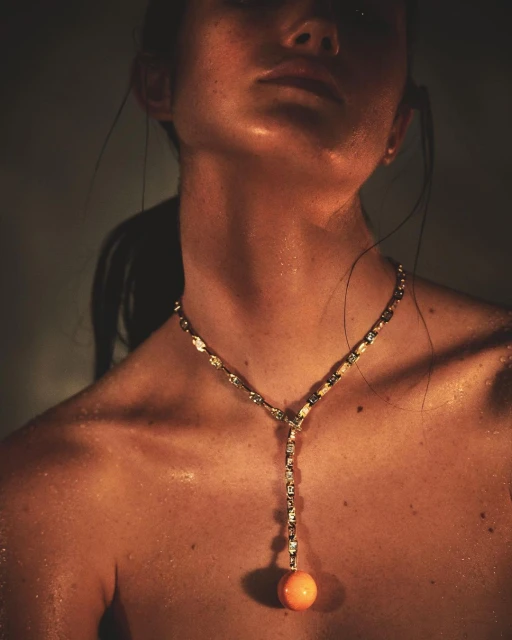 Close-up studio shot of an adult woman with sweaty skin, wearing a jeweled necklace with an orange spherical pendant.