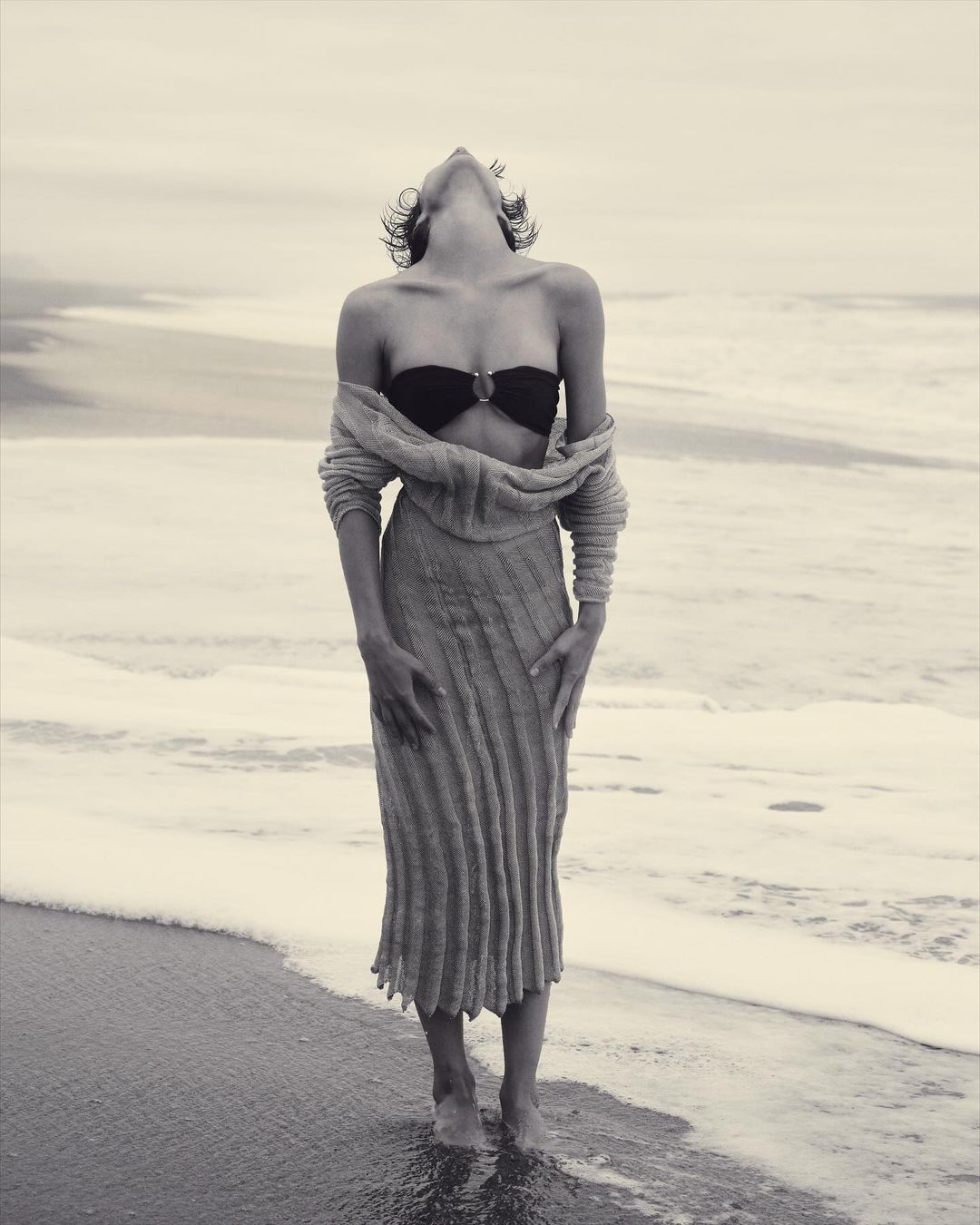 A young adult woman poses in swimwear and a long sweater at the beach, with her head tilted back and ocean waves in the background.