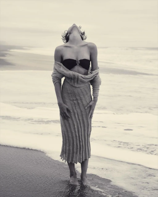 A young adult woman poses in swimwear and a long sweater at the beach, with her head tilted back and ocean waves in the background.
