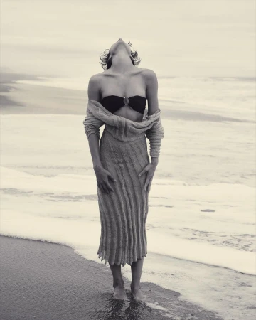 A young adult woman poses in swimwear and a long sweater at the beach, with her head tilted back and ocean waves in the background.