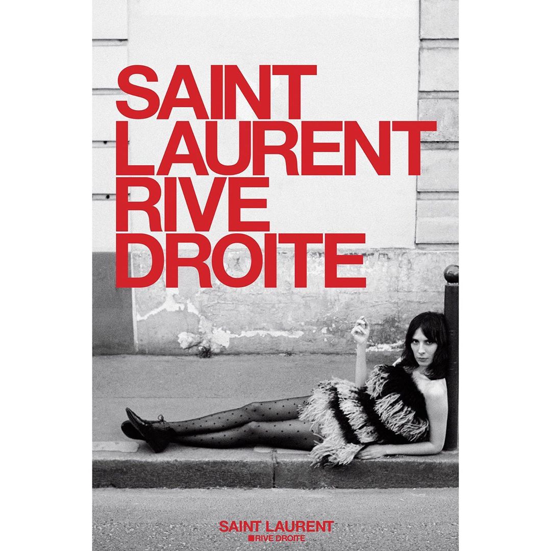 A woman lies on a sidewalk next to a wall with the brand SAINT LAURENT RIVE DROITE in red text above her in a black and white fashion editorial.