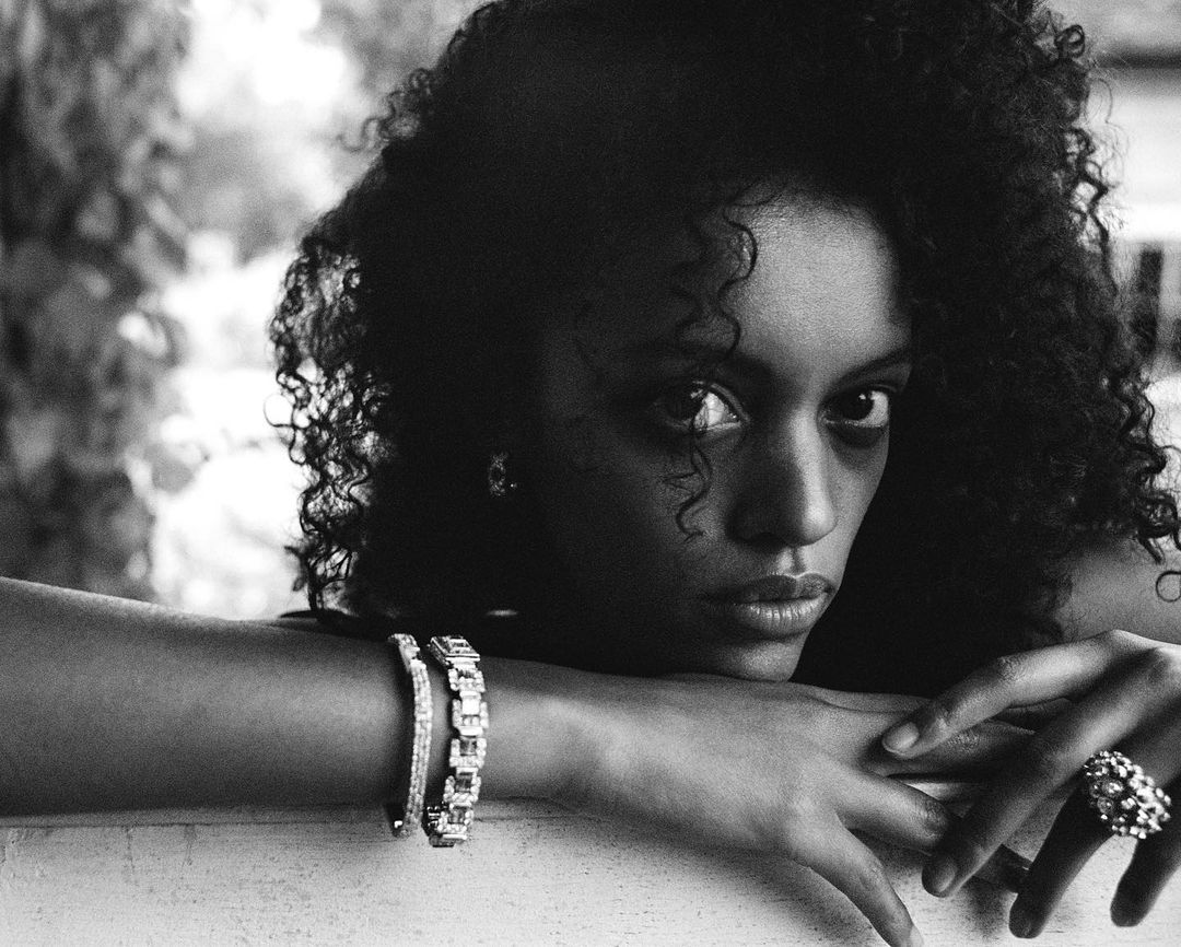 Close-up black and white portrait of a woman with curly hair resting her chin on crossed arms, wearing diamond bracelets and a large ring.
