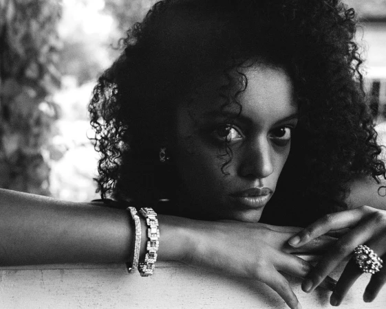 Close-up black and white portrait of a woman with curly hair resting her chin on crossed arms, wearing diamond bracelets and a large ring.