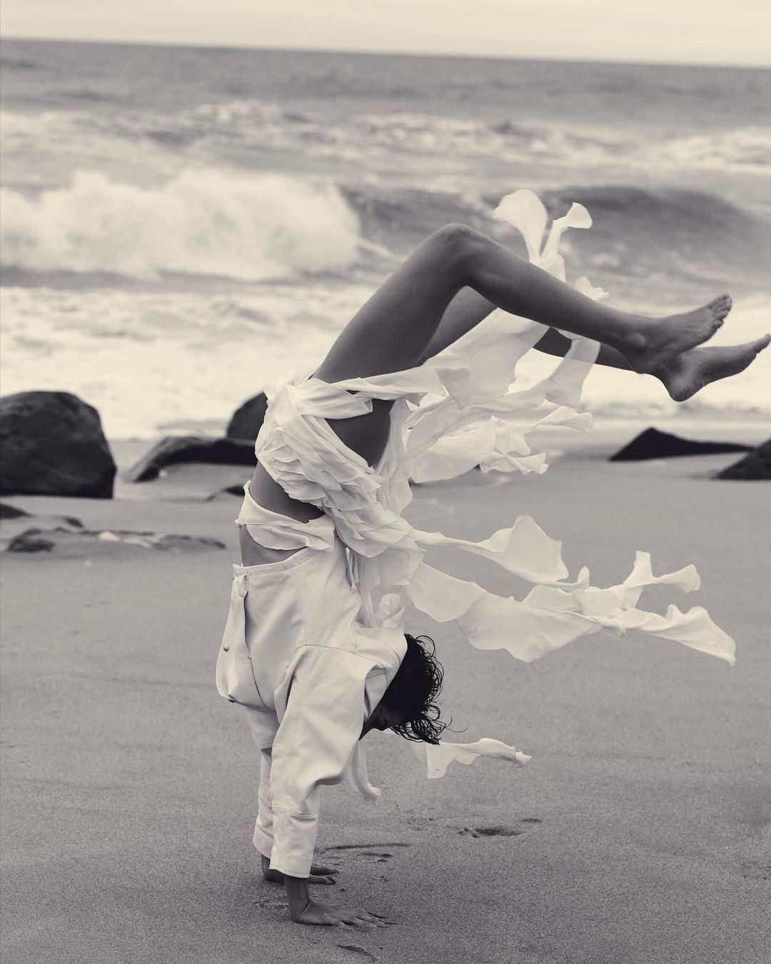Monochrome action shot of a person doing a handstand on a sandy beach wearing tattered white clothing