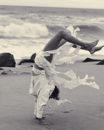 Monochrome action shot of a person doing a handstand on a sandy beach wearing tattered white clothing