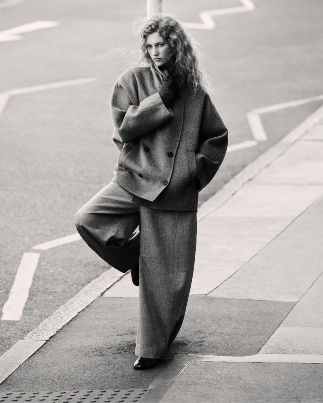 Black and white image of a woman in a suit standing on one leg on a city street with geometric markings, looking directly at the camera.