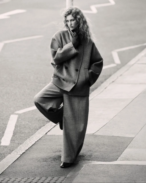 Black and white image of a woman in a suit standing on one leg on a city street with geometric markings, looking directly at the camera.