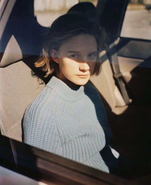 A young woman with short blonde hair sits in the back seat of a car, wearing a light blue sweater, looking toward the camera.