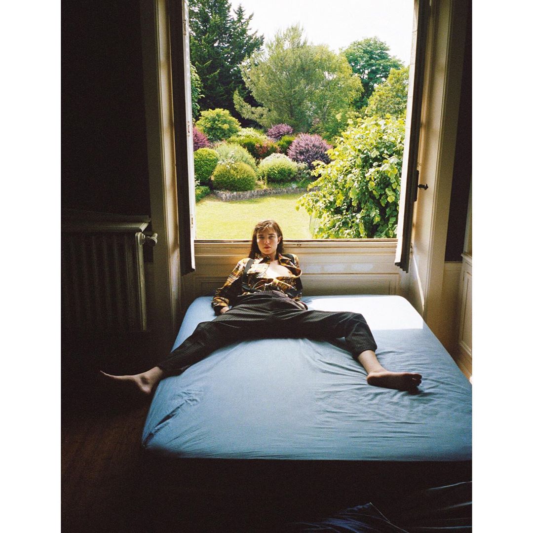 Young woman with brown hair and casual clothing sits cross-legged on a blue bed in front of an open window with a garden view.