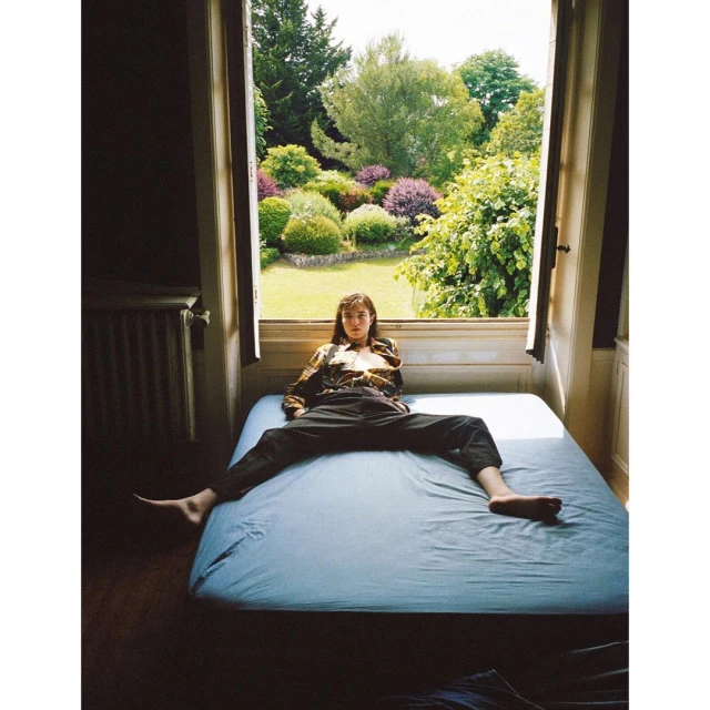Young woman with brown hair and casual clothing sits cross-legged on a blue bed in front of an open window with a garden view.