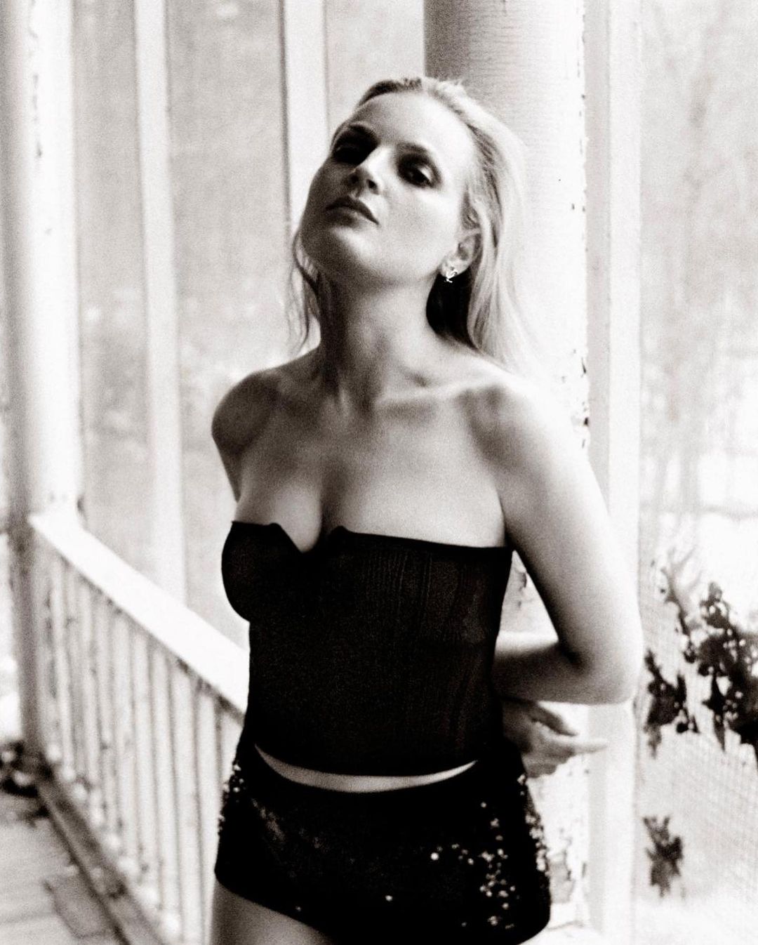 Black and white portrait of a woman with strapless top and shorts, leaning against a weathered column with vegetation in the background.