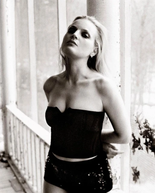 Black and white portrait of a woman with strapless top and shorts, leaning against a weathered column with vegetation in the background.