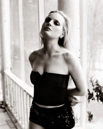 Black and white portrait of a woman with strapless top and shorts, leaning against a weathered column with vegetation in the background.