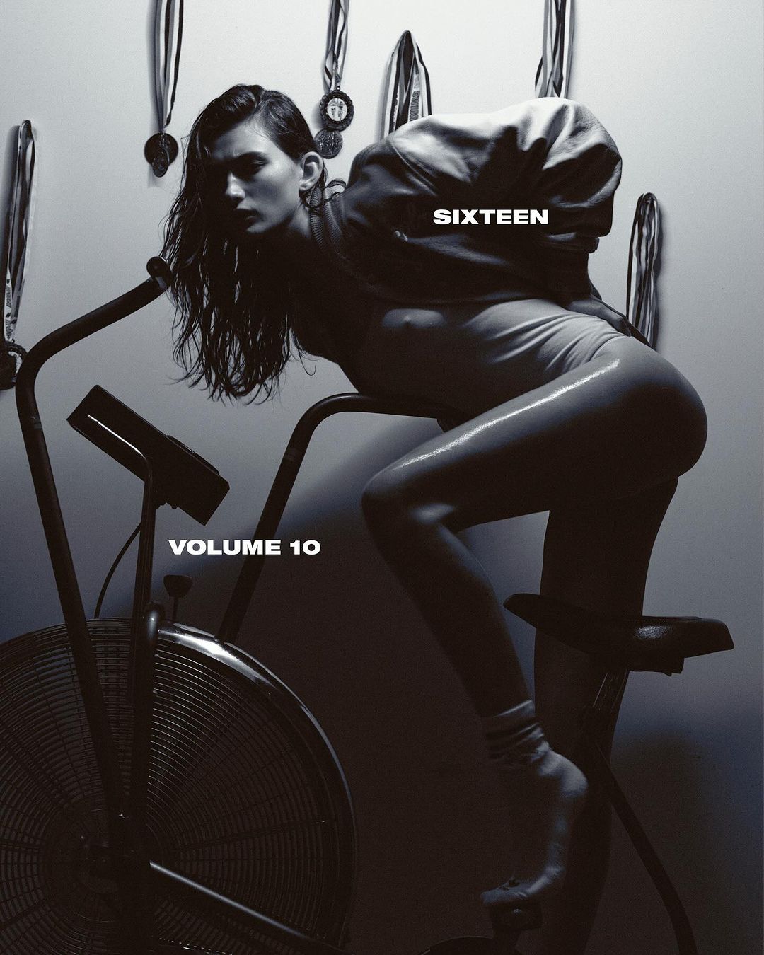 Monochrome image featuring a woman in a bodysuit leaning on an exercise bike with medals hanging in the background. Studio lighting.
