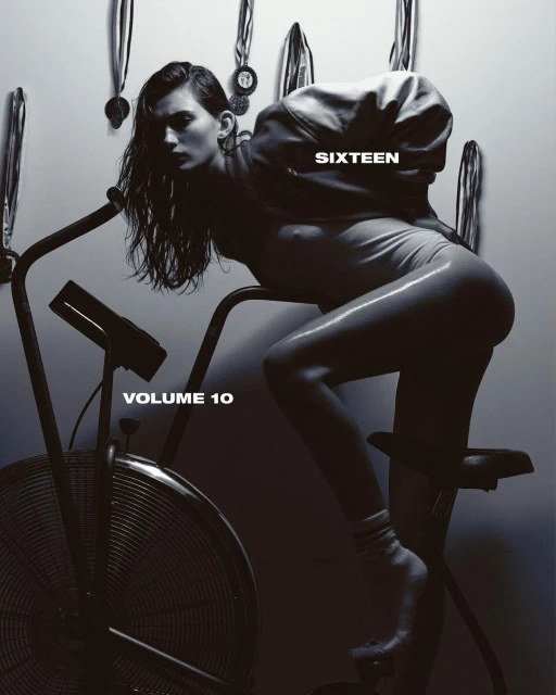 Monochrome image featuring a woman in a bodysuit leaning on an exercise bike with medals hanging in the background. Studio lighting.