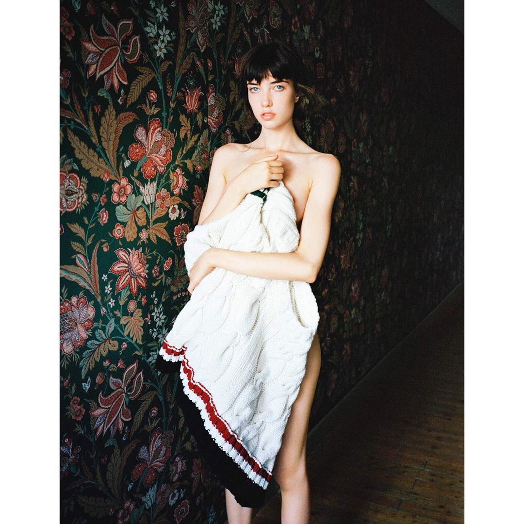 A woman holding a white cable-knit sweater against her chest in front of a dark floral wallpaper background.