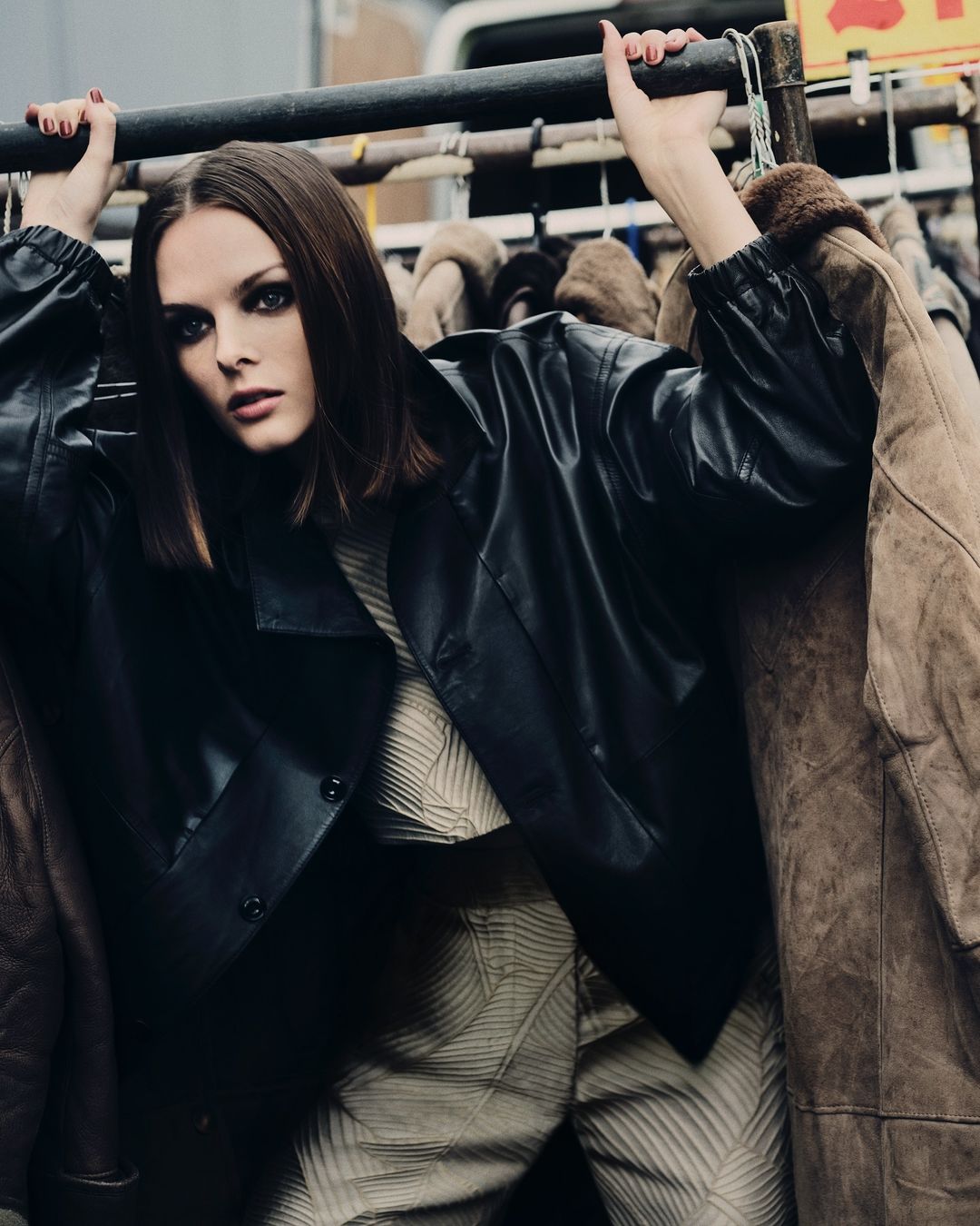 A woman with dark hair and smoky eye makeup is posing among a rack of jackets wearing a leather jacket and patterned top and pants.