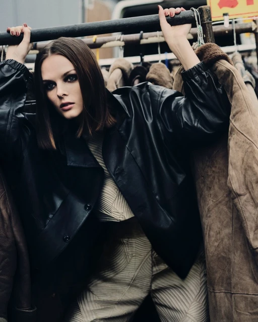 A woman with dark hair and smoky eye makeup is posing among a rack of jackets wearing a leather jacket and patterned top and pants.