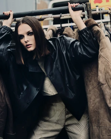 A woman with dark hair and smoky eye makeup is posing among a rack of jackets wearing a leather jacket and patterned top and pants.