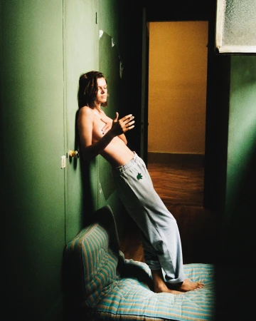 A young, shirtless man leans against a green wall indoors, wearing sweatpants and standing barefoot on a striped mattress.