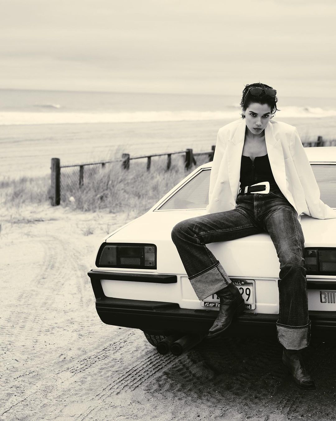 Monochrome image of a woman in a white blazer and jeans sitting on the hood of a car near the beach on a cloudy day.