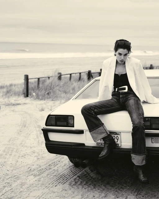 Monochrome image of a woman in a white blazer and jeans sitting on the hood of a car near the beach on a cloudy day.