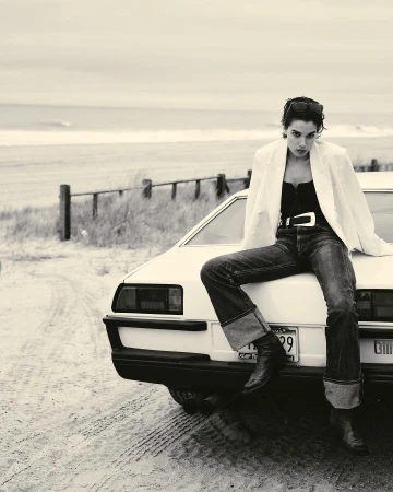 Monochrome image of a woman in a white blazer and jeans sitting on the hood of a car near the beach on a cloudy day.