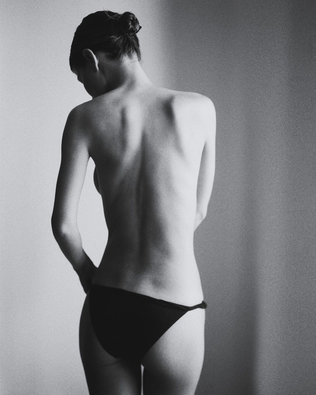 Black and white shot of a woman with hair in a bun, back facing the camera, wearing bikini bottoms in a studio setting.