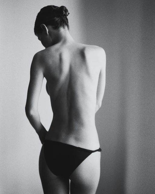 Black and white shot of a woman with hair in a bun, back facing the camera, wearing bikini bottoms in a studio setting.