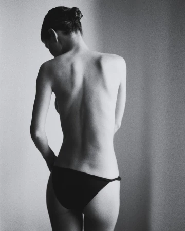 Black and white shot of a woman with hair in a bun, back facing the camera, wearing bikini bottoms in a studio setting.