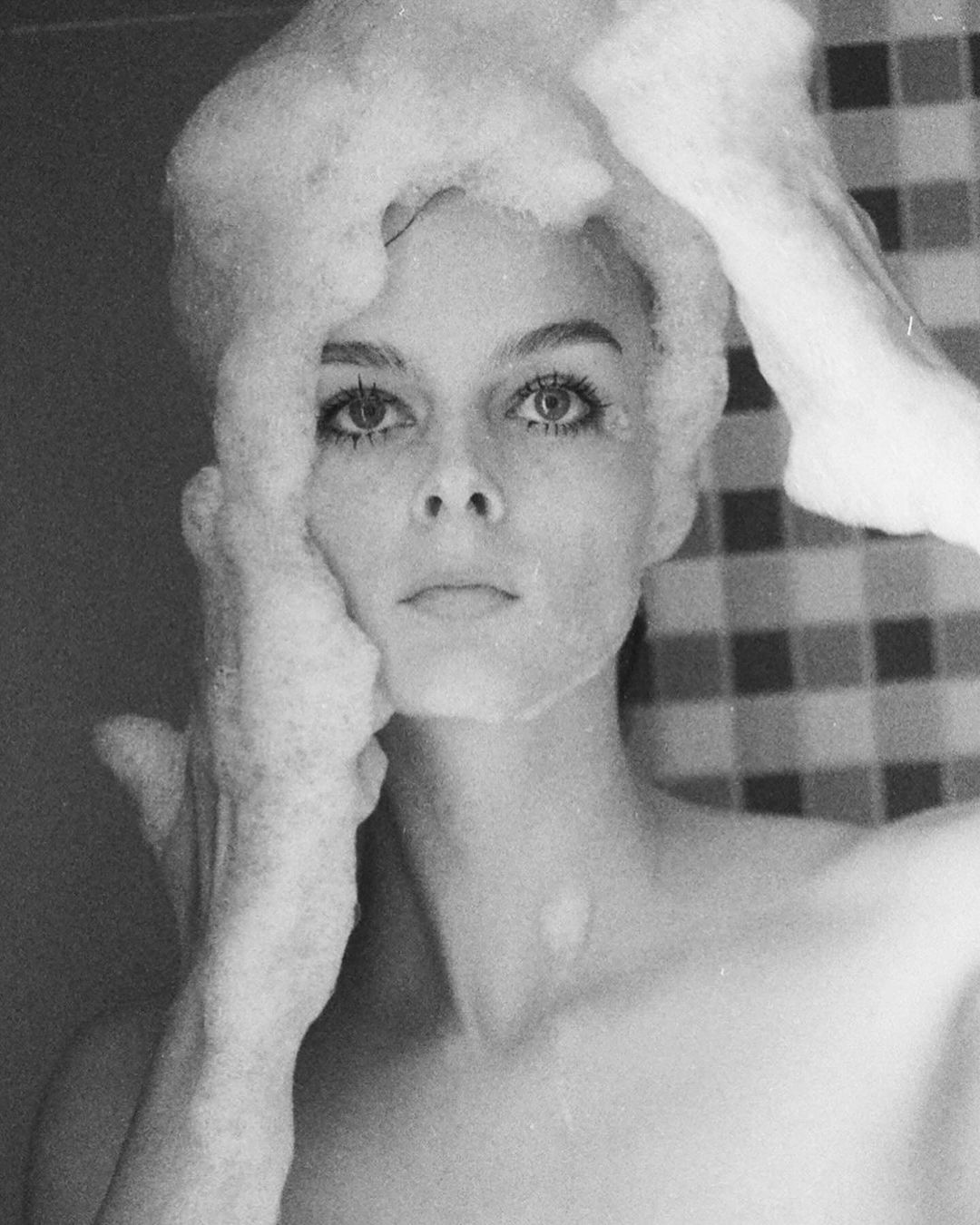 Black and white portrait of a young woman in a bathroom shower, covered in bath soap foam and looking directly at the camera.